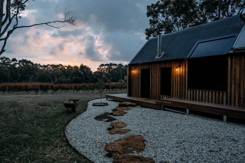 A modern wooden cabin set in a rural landscape at twilight, with a gravel path leading to it.